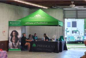 Paramount health plan booth at the Lucas County Fair, featuring a green canopy with staff seated at a table providing information. A large banner shows smiling individuals, and a mobile unit is visible in the background