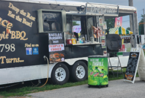 Food truck vendor named Dre’s Place at the Lucas County Fair, featuring a black trailer with BBQ signage, an order window with displayed menu items, a grill setup outside, and a Mountain Dew advertisement in front.